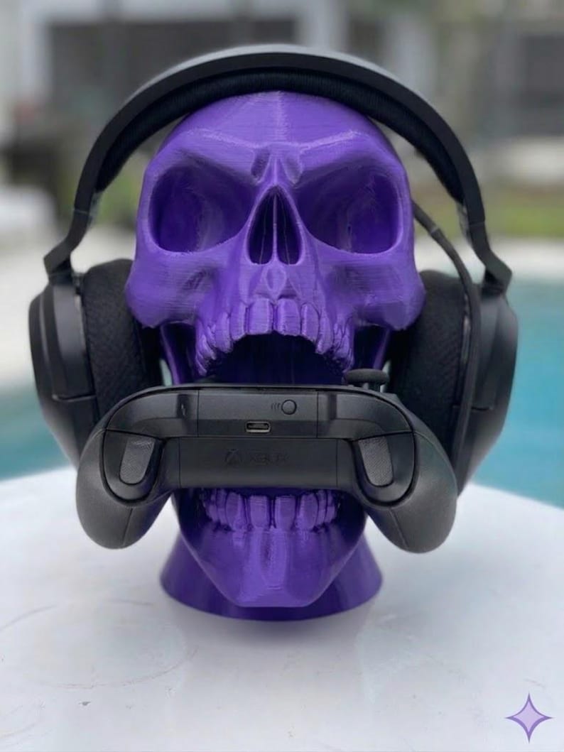May include: A purple 3D printed skull display stand holding a pair of black over-ear gaming headphones on its cranium and a black wireless gaming controller positioned horizontally within its open jaw, showcasing a matte finish and textured surface.