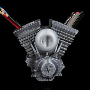 May include: A silver-toned V-twin engine shaped desk organizer with a weathered metallic finish. The central circular cover features the embossed text V2. Two hollow cylinder heads serve as holders for various pens and colored pencils.