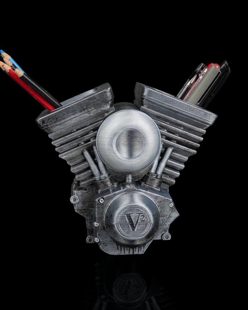 May include: A silver-toned V-twin engine shaped desk organizer with a weathered metallic finish. The central circular cover features the embossed text V2. Two hollow cylinder heads serve as holders for various pens and colored pencils.