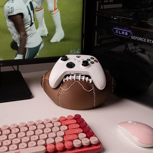 Op de afbeelding: Een witte draadloze gamecontroller rustend op een bruine, getextureerde standaard in de vorm van een American football met witte stiksels. De controller heeft zwarte analoge sticks, een D-pad en veelkleurige actieknoppen voor een computermonitor.