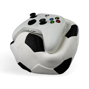 Op de afbeelding: Een witte draadloze Xbox-controller rustend op een pluche, zacht kussen in de vorm van een voetbal. De controller is voorzien van zwarte analoge sticks, een zwarte D-pad en de standaard gekleurde A-, B-, X- en Y-knoppen. De voetbal heeft een klassiek zwart-wit patroon.