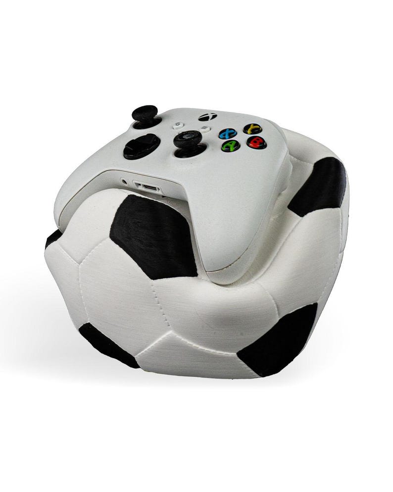 Op de afbeelding: Een witte draadloze Xbox-controller rustend op een pluche, zacht kussen in de vorm van een voetbal. De controller is voorzien van zwarte analoge sticks, een zwarte D-pad en de standaard gekleurde A-, B-, X- en Y-knoppen. De voetbal heeft een klassiek zwart-wit patroon.