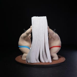 May include: A napkin holder featuring two sumo wrestler figurines standing on a circular brown base. One wrestler wears a blue mawashi and the other wears a red mawashi. They are positioned facing each other, holding a stack of white paper napkins.