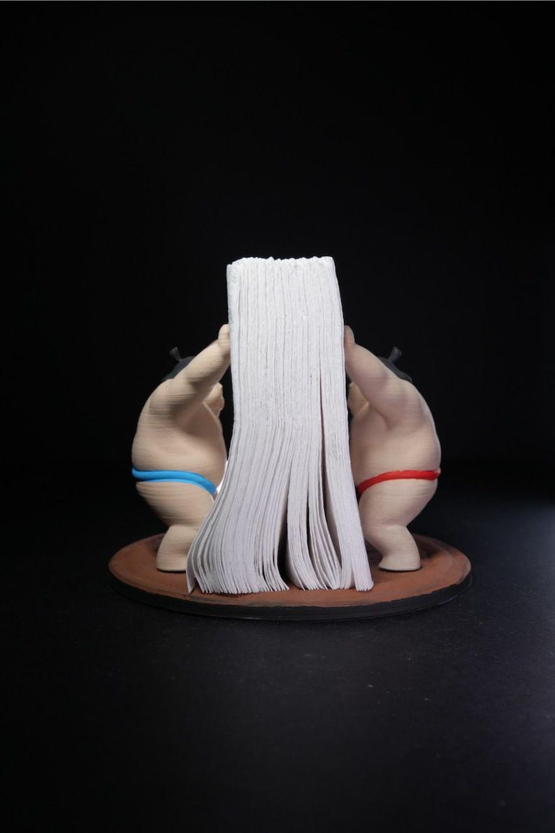 May include: A napkin holder featuring two sumo wrestler figurines standing on a circular brown base. One wrestler wears a blue mawashi and the other wears a red mawashi. They are positioned facing each other, holding a stack of white paper napkins.