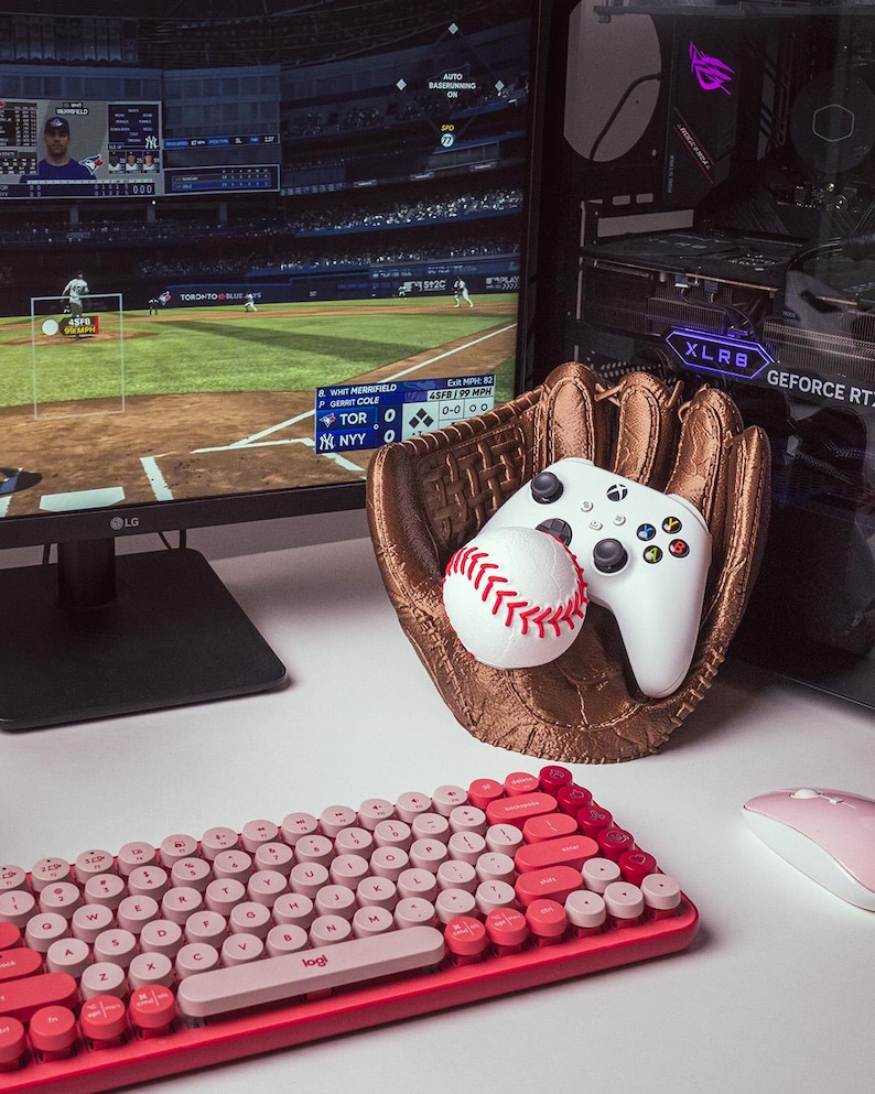 Puede incluir: Un soporte con forma de guante de b&eacute;isbol color bronce que contiene un mando de juego blanco y una pelota de b&eacute;isbol blanca con costuras rojas, junto a un monitor de ordenador con un partido de b&eacute;isbol y un teclado y rat&oacute;n rosas.