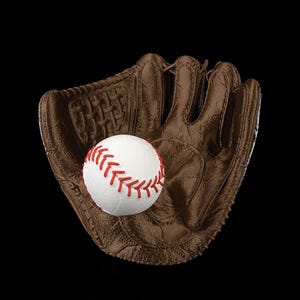 Puede incluir: Un guante de b&eacute;isbol de cuero marr&oacute;n con un dise&ntilde;o de red tejida y costuras visibles, que sostiene una pelota de b&eacute;isbol blanca con cordones rojos prominentes. El guante tiene una superficie texturizada y un bolsillo profundo.