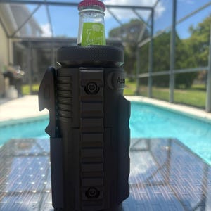 Pode incluir: Um refrigerador de garrafas cil&iacute;ndrico preto fosco com ader&ecirc;ncia texturizada, um sistema de trilho vertical com dois parafusos vis&iacute;veis e um abridor de garrafas integrado na lateral. Uma garrafa de vidro com tampa vermelha est&aacute; inserida.