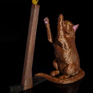 May include: A brown cat figurine with green eyes and pink inner ears reaching up with a paw toward a small yellow bird perched on top of a brown wooden-textured axe handle. The axe head is black and rests on the ground beside the cat.
