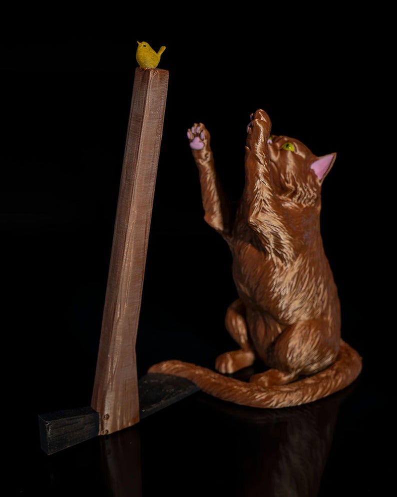 May include: A brown cat figurine with green eyes and pink inner ears reaching up with a paw toward a small yellow bird perched on top of a brown wooden-textured axe handle. The axe head is black and rests on the ground beside the cat.