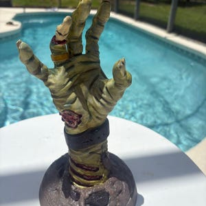 May include: A sculpted, upright zombie hand figurine featuring weathered, pale green skin with visible muscle texture, a dark wrist cuff, a gold-colored ring on the ring finger, and a small insect attached to the forearm, mounted on a circular base.