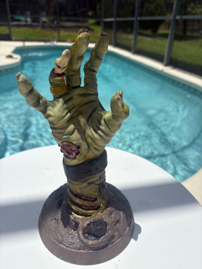 May include: A sculpted, upright zombie hand figurine featuring weathered, pale green skin with visible muscle texture, a dark wrist cuff, a gold-colored ring on the ring finger, and a small insect attached to the forearm, mounted on a circular base.