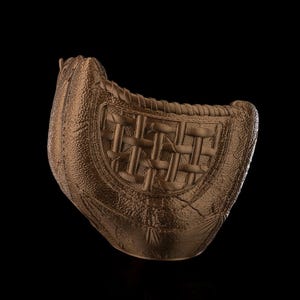 Puede incluir: Un guante de b&eacute;isbol de color bronce con una superficie texturizada similar al cuero y un patr&oacute;n de red trenzada. El guante presenta un acabado met&aacute;lico s&oacute;lido con detalles de costuras visibles a lo largo de los bordes y una forma curva.