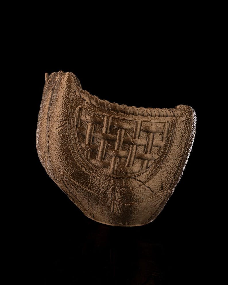 Puede incluir: Un guante de b&eacute;isbol de color bronce con una superficie texturizada similar al cuero y un patr&oacute;n de red trenzada. El guante presenta un acabado met&aacute;lico s&oacute;lido con detalles de costuras visibles a lo largo de los bordes y una forma curva.