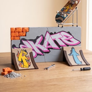 Puede incluir: Un set de fingerboard que incluye un monopatín en miniatura con la marca Pro Artisan, un muro gris estilo hormigón con el texto SKATE en grafiti rosa, dos rampas pequeñas con gráficos, una llave metálica, un destornillador y ladrillos.