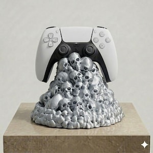 Op de afbeelding: Een wit en zwart videogame-controller rustend op een gebeeldhouwde basis bestaande uit een dichte stapel metallic zilveren schedels. De controller is voorzien van standaard richtingsknoppen, analoge sticks en actiesymbolen op de voorzijde.