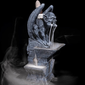 Puede incluir: Un quemador de incienso con forma de g&aacute;rgola de textura de piedra gris oscuro que presenta una criatura alada sobre un pedestal escalonado. Un cono de incienso encendido libera humo blanco que fluye desde la boca hacia un recipiente.