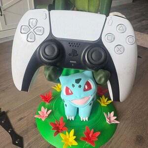 Op de afbeelding: Een wit en zwart videogame-controller rustend op een 3D-geprinte standaard in de vorm van het personage Bulbasaur. De standaard heeft een groene ronde basis versierd met kleine rode, roze en gele bloemen en groene ranken.