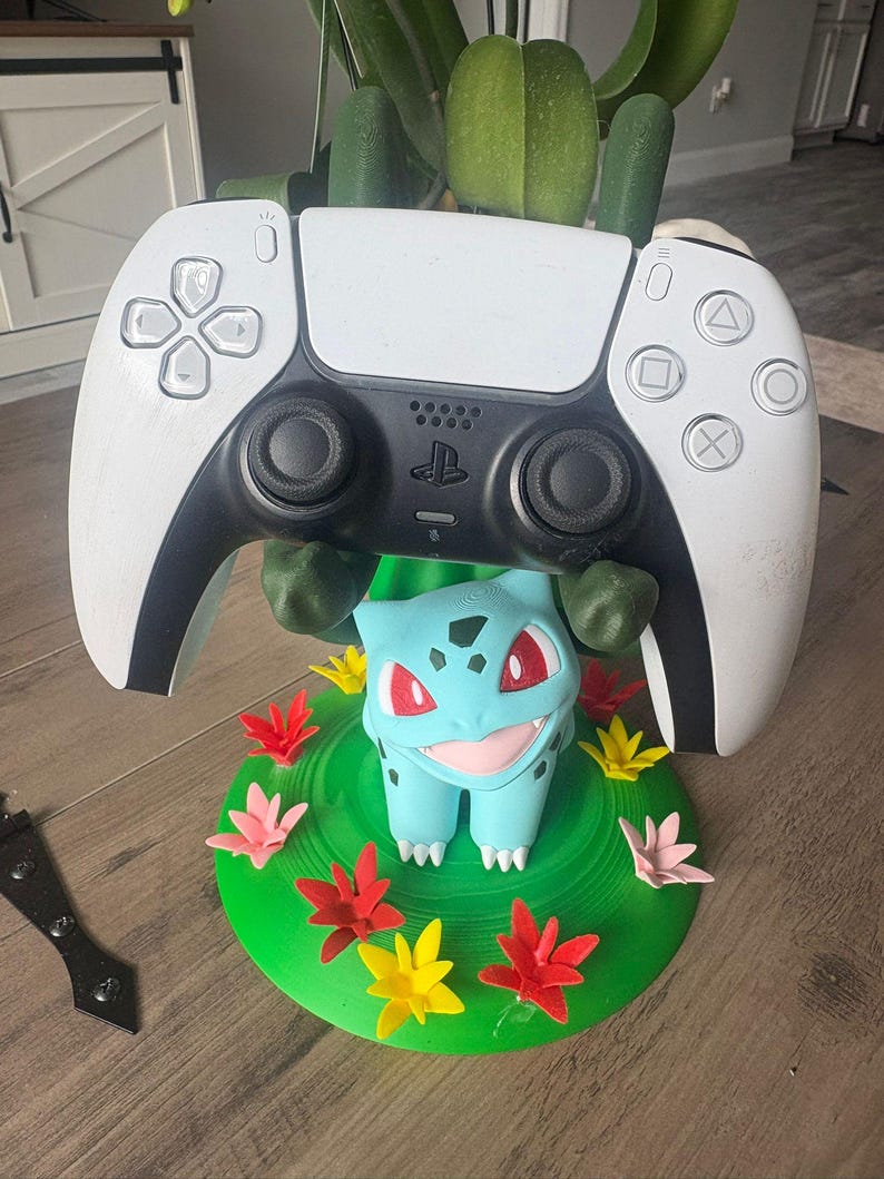 Op de afbeelding: Een wit en zwart videogame-controller rustend op een 3D-geprinte standaard in de vorm van het personage Bulbasaur. De standaard heeft een groene ronde basis versierd met kleine rode, roze en gele bloemen en groene ranken.