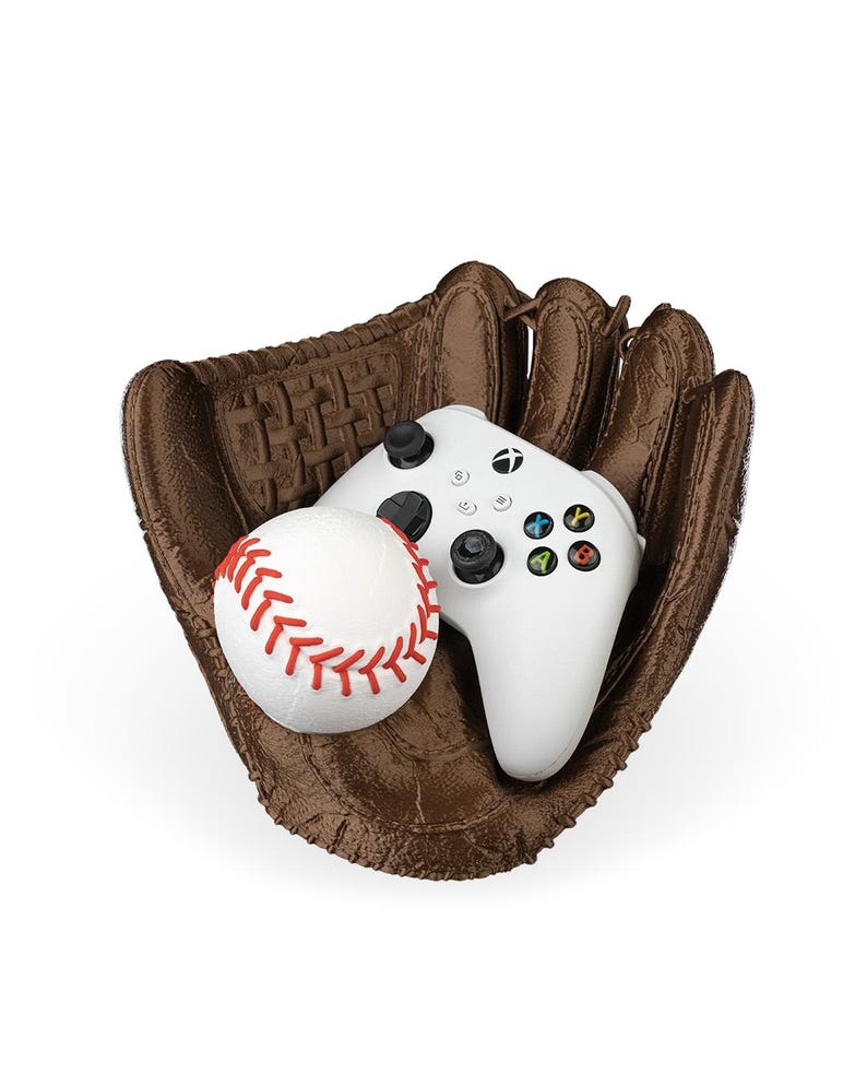 Puede incluir: Un guante de b&eacute;isbol marr&oacute;n texturizado que sostiene una pelota de b&eacute;isbol blanca con costuras rojas y un mando de videojuego inal&aacute;mbrico blanco con joysticks negros, una cruceta y cuatro botones de acci&oacute;n de colores A, B, X e Y.