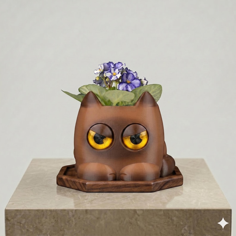 May include: A brown, cat-shaped planter with large, glossy yellow eyes and pointed ears. The planter sits on a dark brown, hexagonal wooden base and contains a small green plant with purple flowers blooming from the top of the cat's head.