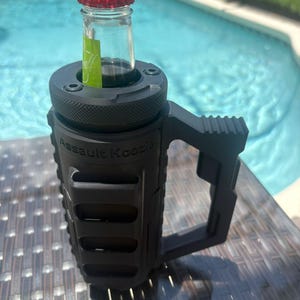 Pode incluir: Um porta-bebidas preto fosco com um exterior texturizado de estilo t&aacute;tico e uma al&ccedil;a lateral resistente. As palavras Assault Koozie est&atilde;o gravadas na frente. Uma garrafa de vidro com tampa vermelha est&aacute; inserida na abertura superior.