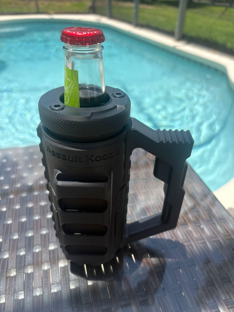 Pode incluir: Um porta-bebidas preto fosco com um exterior texturizado de estilo t&aacute;tico e uma al&ccedil;a lateral resistente. As palavras Assault Koozie est&atilde;o gravadas na frente. Uma garrafa de vidro com tampa vermelha est&aacute; inserida na abertura superior.