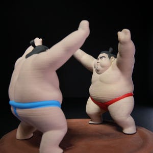 May include: Two sumo wrestler figurines standing on a circular brown base. One figure wears a bright blue mawashi and faces away, while the other wears a red mawashi and faces forward with arms raised. Both figures have black hair tied in topknots.