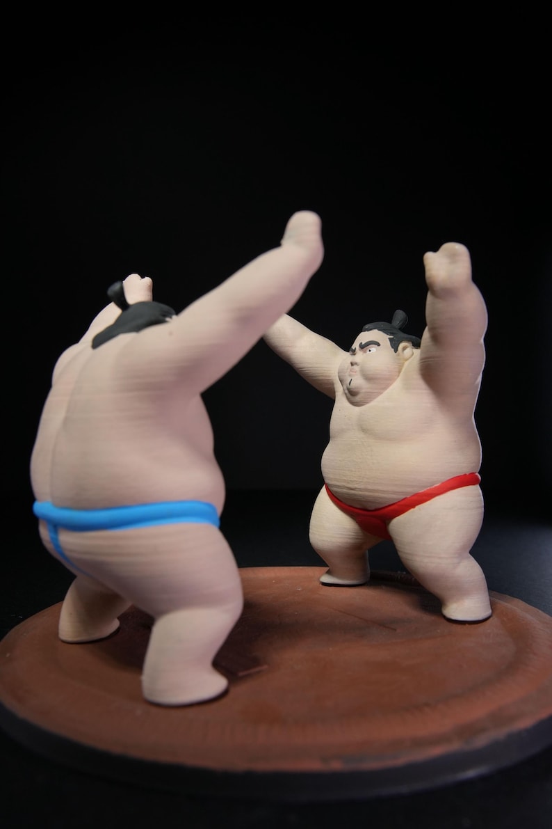 May include: Two sumo wrestler figurines standing on a circular brown base. One figure wears a bright blue mawashi and faces away, while the other wears a red mawashi and faces forward with arms raised. Both figures have black hair tied in topknots.