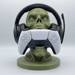 May include: An olive green skull-shaped stand holding a pair of black over-ear gaming headphones on its head and a white and black wireless game controller in its mouth. The stand features a circular base and detailed skeletal facial features.