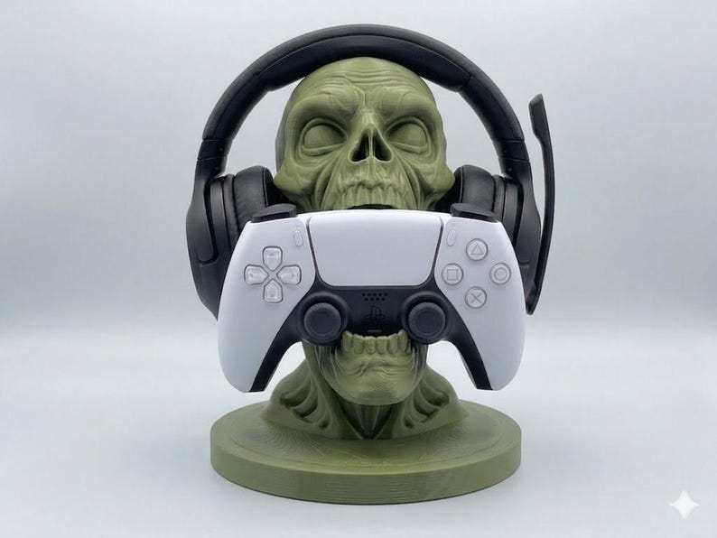 May include: An olive green skull-shaped stand holding a pair of black over-ear gaming headphones on its head and a white and black wireless game controller in its mouth. The stand features a circular base and detailed skeletal facial features.