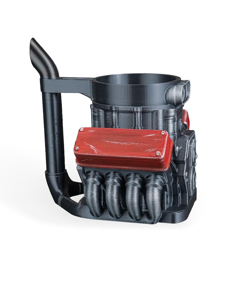 May include: A black mug shaped like a V8 engine block with a tall exhaust pipe handle. The design features a prominent red valve cover with visible screw details and metallic silver exhaust headers extending from the side of the engine block structure.