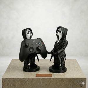 May include: Two black figurines with white ghost masks and hooded robes. One figurine holds a black video game controller with coloured buttons, while the other stands with empty hands outstretched. Both figures are mounted on circular black bases.