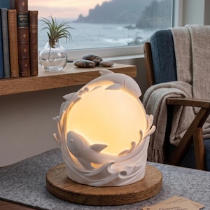 Op de afbeelding: Een witte keramische lamp met een cirkelvormig gloeiend licht omringd door gebeeldhouwde golven en twee vissen. De lamp staat op een ronde houten voet. Een klein beige kaartje met de tekst Oceanic Illumination ligt op het grijze oppervlak.