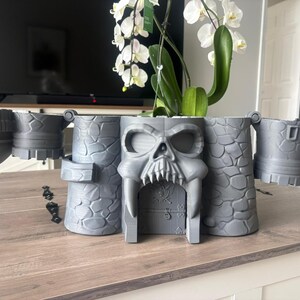 Op de afbeelding: Een grijze 3D-geprinte plantenbak in kasteelthema met een opvallend schedelontwerp bij de centrale ingang. De structuur bevat muren met een steenachtige textuur, zijtorens en scharnierende delen die naar buiten uitsteken voor een brede uitstraling.