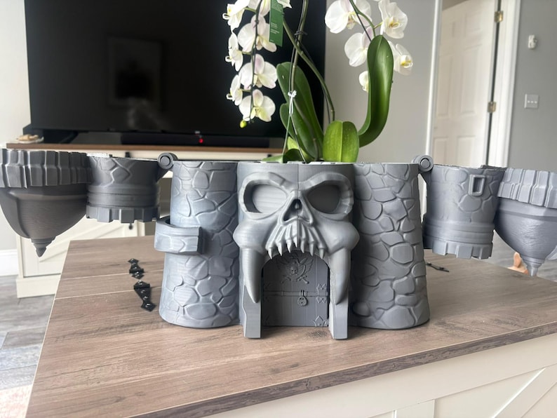 Op de afbeelding: Een grijze 3D-geprinte plantenbak in kasteelthema met een opvallend schedelontwerp bij de centrale ingang. De structuur bevat muren met een steenachtige textuur, zijtorens en scharnierende delen die naar buiten uitsteken voor een brede uitstraling.