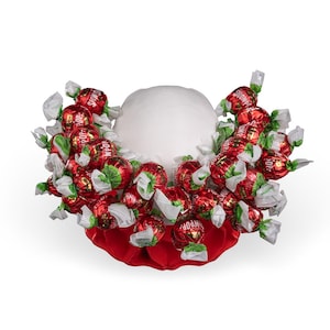 May include: A white foam sphere base adorned with numerous red foil-wrapped sweets, each featuring white twisted ends and the text "SHOWPOP" printed in white. The sweets are arranged in a dense cluster around the centre of the sphere.