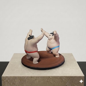 May include: A figurine featuring two sumo wrestlers in a grappling stance on a circular brown platform. One wrestler wears a red mawashi, while the other wears a blue mawashi. Both figures have black hair styled in a topknot and are facing each other.