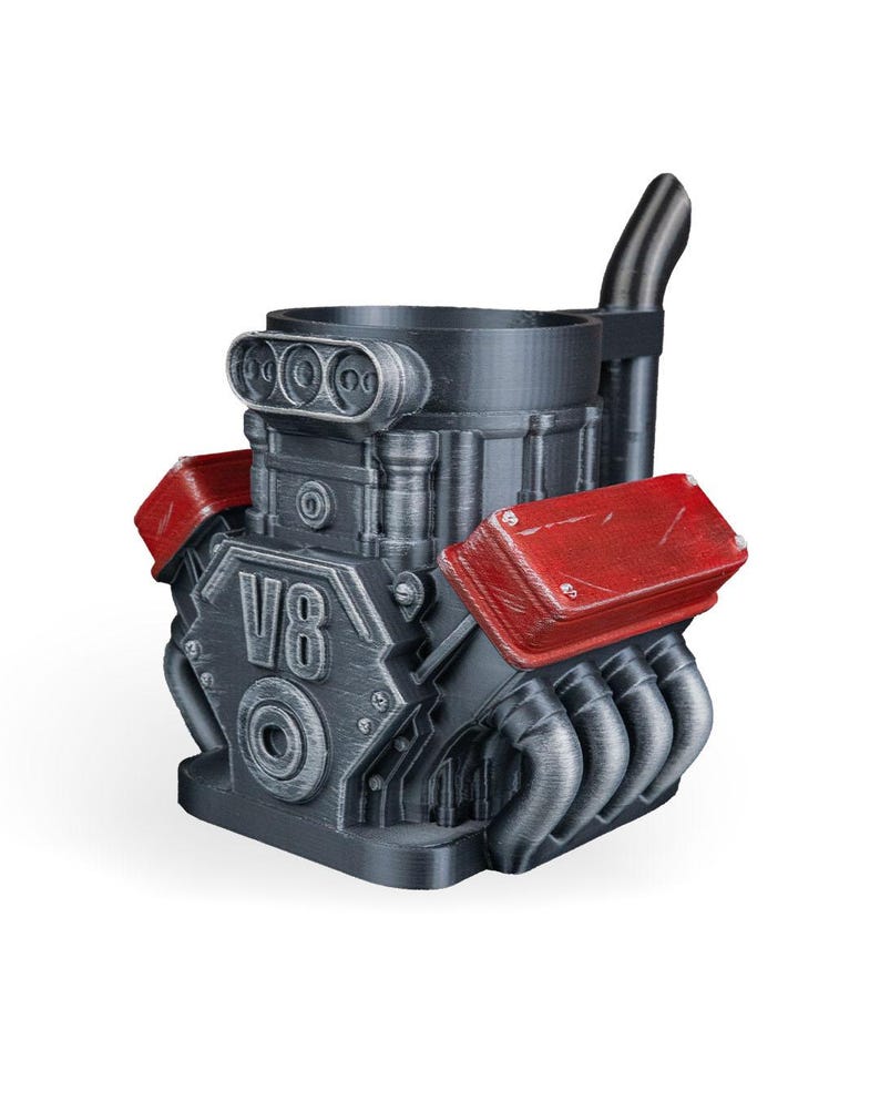 May include: A dark gray mug shaped like a V8 engine block with red valve covers and a black exhaust pipe extending from the back. The front of the engine block features the embossed text V8, along with detailed mechanical components and pipes.