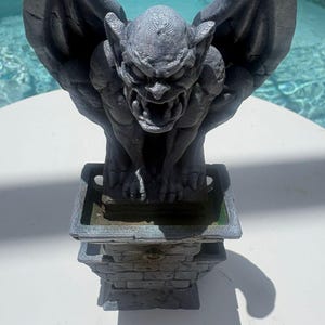 Puede incluir: Una estatua de g&aacute;rgola gris con alas grandes y expresi&oacute;n amenazante, posada sobre un pedestal cuadrado con textura de ladrillo. La figura presenta una definici&oacute;n muscular detallada y una superficie texturizada sobre una base.