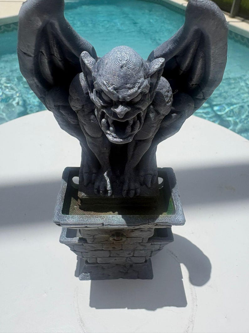 Puede incluir: Una estatua de g&aacute;rgola gris con alas grandes y expresi&oacute;n amenazante, posada sobre un pedestal cuadrado con textura de ladrillo. La figura presenta una definici&oacute;n muscular detallada y una superficie texturizada sobre una base.