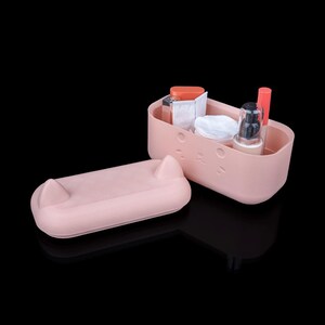 May include: A light pink rectangular storage container with a matching lid featuring two small pointed ears. The front of the container has a subtle embossed cat face design. Inside are various cosmetic items including a pump bottle and tubes.