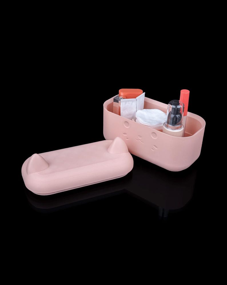 May include: A light pink rectangular storage container with a matching lid featuring two small pointed ears. The front of the container has a subtle embossed cat face design. Inside are various cosmetic items including a pump bottle and tubes.