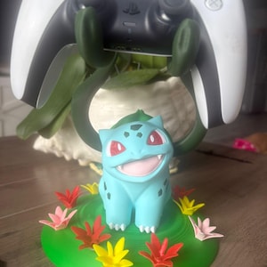 Op de afbeelding: Een lichtblauw Bulbasaur-figuur op een ronde groene basis versierd met kleine rode, gele en roze bloemen. Het figuur heeft twee donkergroene, rankachtige armen die omhoog reiken om een wit-zwarte videospelcontroller vast te houden.