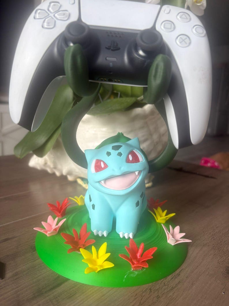 Op de afbeelding: Een lichtblauw Bulbasaur-figuur op een ronde groene basis versierd met kleine rode, gele en roze bloemen. Het figuur heeft twee donkergroene, rankachtige armen die omhoog reiken om een wit-zwarte videospelcontroller vast te houden.