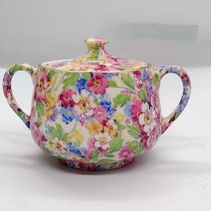 May include: A ceramic sugar bowl with a lid and two side handles, featuring a colourful floral pattern with pink, yellow, blue, and green blossoms on a cream-coloured background. The bowl has a rounded body and a matching lid with a small knob handle.