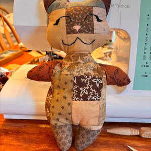 May include: A stuffed cat toy constructed from various brown, tan, and cream fabric patches. The cat features embroidered black eyes and a mouth, a small pink stitched nose, and pointed ears. The patchwork design includes floral, star, and solid patterns.