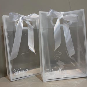 May include: Two clear rectangular gift bags with white trim and white satin ribbon bows tied at the top handles. Each bag features a custom name printed in white script lettering on the front, specifically the names Freya and Hadley.