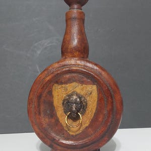 Könnte beinhalten: Eine braune, lederbezogene Karaffe mit kugelförmigem Verschluss. Die Vorderseite zeigt eine mittige, schildförmige Einlage in Beige mit Blumenprägung, in deren Mitte sich eine dunkle Metall-Löwenkopfskulptur mit einem goldenen Ring befindet.