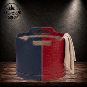May include: A cylindrical storage basket featuring a split design with smooth navy blue leather on one side and red crocodile-embossed leather on the other. It includes integrated handles and a beige fabric throw draped over the side.