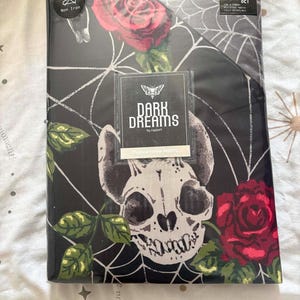 Op de afbeelding: Een zwart king-size dekbedovertrekset in plastic verpakking met een wit spinnenwebpatroon, rode rozen met groene bladeren en een schedelafbeelding. Het label vermeldt Dark Dreams by Rapport, Mystical Forces Bedding en Non Iron.